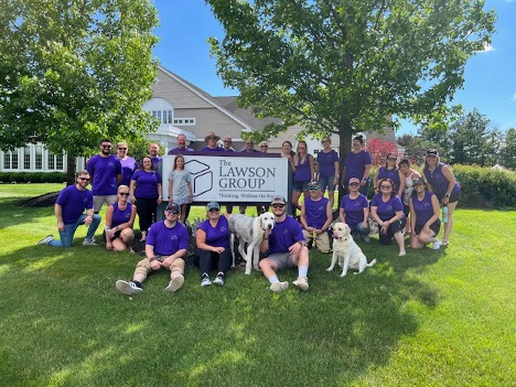 The Lawson Group team poses outside on a sunny day in front of the company’s Concord, NH office. Everyone is wearing matching purple shirts, gathered around the company sign that reads “The Lawson Group – Thinking Without the Box.” Three friendly dogs sit with the group on the green lawn, surrounded by trees and bright blue sky.